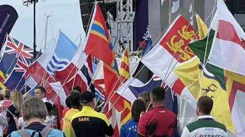 People in multi-coloured tracksuits hold up large multi-coloured flags on white flag poles.