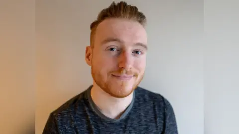 Eoin Reid A man with short ginger hair and ginger stubble. He is smiling and looking at the camera. He is wearing a dark grey top.