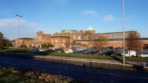 Brick building with green roof. Some of the roof is pointed and other parts are like a castle's battlements. There are cars parked in front and a road. There are bushes and grass. 
