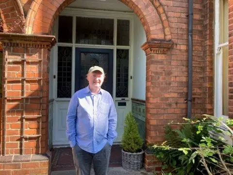 BBC / Sally Fairfax Stephen Ingram is smiling. He wears a blue shirt, grey trousers and grey cap and is standing in front of the door of a red-brick house