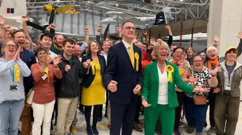 A crowd of people wearing Liberal Democrat rosettes cheer and celebrate. 