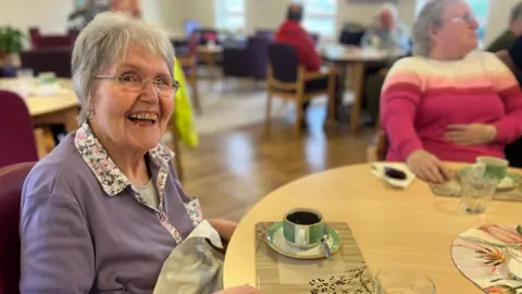 An older lady with short grey hair and glasses wearing a lavender blouse sits at a table with a cup of coffee and smiles at the camera.