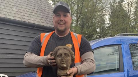 Digger driver Craig Crawley, wearing a dark top and orange high vis vest, holding the roman lady head. It is covered in earth.
