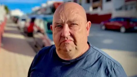 Billy George is standing in the middle of a road in view of the camera. He is bald and wearing a Navy Blue T-shirt. The sun is producing shadows from cars and houses that line both sides of the road behind it. The background is blurred.