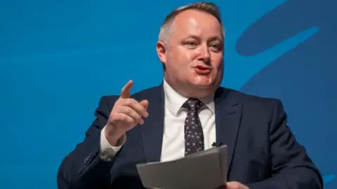 Darren Millar is delivering a speech to an audience. His right finger is pointing upwards. He is wearing a suit, tie and shirt, and is stood before a dark background.