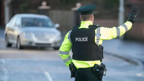 A generic photograph of a policeman dressed in high visibility yellow jacket and black vest with the word police written in silver capital letters on the back. He is wearing black gloves and raising his arm to stop a silver car which is approaching on the road.