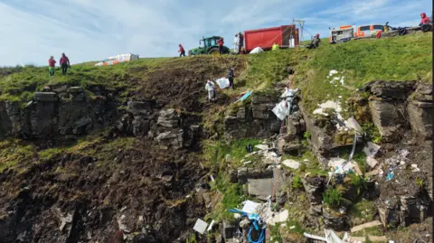 Central Beacons Mountain Rescue Team Close up drone image showing people cleaning up piles of rubbish on the side of a mountain. The volunteers are attached to ropes, with crews supporting them on the top of the hill.