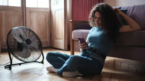 A young woman scrolls on her phone as she sits on the wooden floor of her house in front of a free standing black fan.