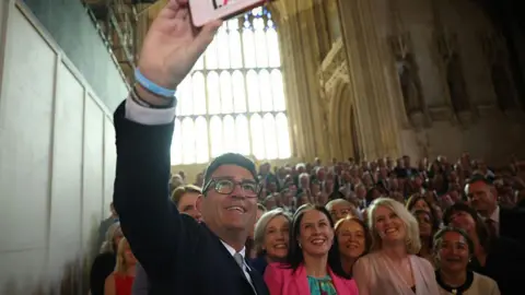 Andy Burnham wearing a black suit, white shirt, and grey tie taking a selfie of a large crowd of Labour MPs in Westminster on the day he is sworn in as an MP