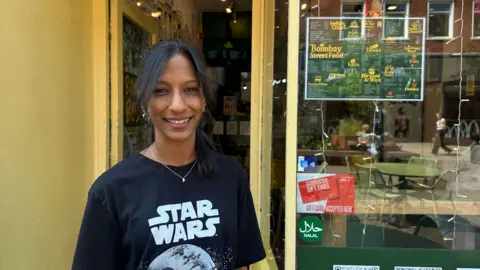 A woman wearing a black star wars shirt and a silver necklace. She is standing outside her restaurant.
