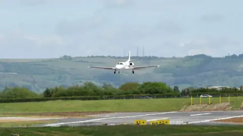 Gloucestershire Airport Small white plane landing on a runway at Gloucestershire Airport surrounded by grass and hills.