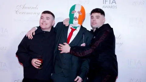 Reuters Three men are standing smiling for a photo. Left, is wearing a black coat and holding a glass. He is smiling at the camera and has short brown hair. The one in the middle is wearing a grey tartan suit and a red tie. He is also wearing a balaclava which is green white and orange striped. On the right, a man has his arms around the one in the middle. He is wearing a dark coloured jacket and has short black hair.