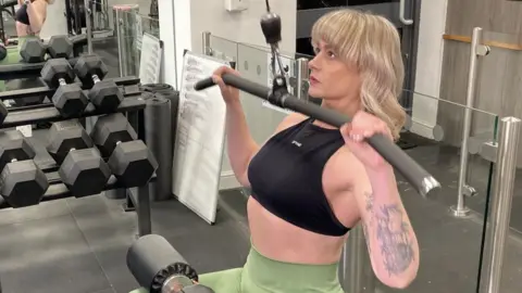 Georgina Cox working out with a pull-down bar in a gym. She is wearing green leggings and a black top. She is in the process of pulling the bar down towards her chest.