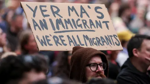Reuters A person holds a placard saying "Yer ma's an immigrant. We're all immigrants uring a 'United Against Racism' rally after days of anti-immigration violence, following a knife attack on June 8, which left a man seriously injured and prompted police to declare a critical incident, in Belfast, Northern Ireland, June 13, 2026. REUTERS/Isabel Infantes