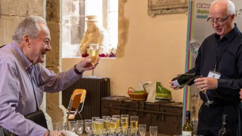 Dale Hodgetts A man holding up a flute in a cheers motion to the man in front of him, who is pouring wine into another flute. They are stood either side of a table which is filled with flutes of alcohol.