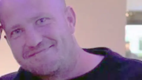 Derek Russell, a shaven headed man wearing a black top, smiles for the camera while standing indoors. 