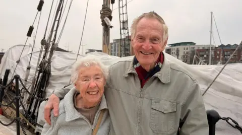 Peter has his arm around Pat and they both look incredibly happy. A boat is behind them and you can see other buildings on the other side of the dock.