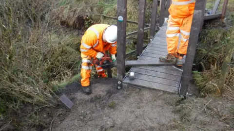 Staffordshire County Council Two workers in orange outfits and wearing white helmets work on a footbridge. One is standing on a boardwalk panel while the other is leaning over and cutting the other end of it with a circular saw.