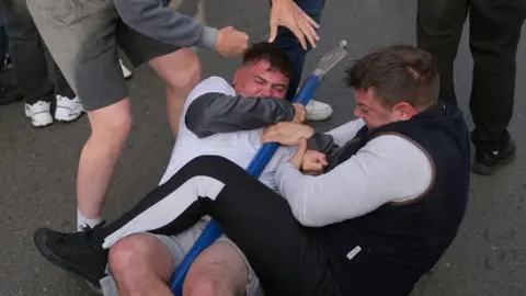 PA Media Two men grappling on the ground trying to keep hold of a blue pole.