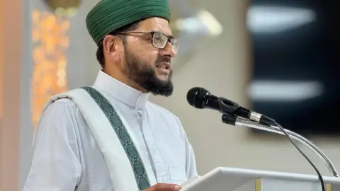 Qari Asim An imam stood at a Minbar giving a sermon. He's dressed in white with a green hat.