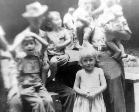 Nancy Barr/MediaPunch via Alamy Dolly Parton, aged 5, with her family 1951 in a black and white image. She stands in a sleeveless white dress while adults holding children stand out of focus behind her. 