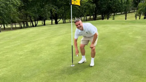 Matthew Vigurs Vigurs wearing a white polo top and beige shorts bends over and picks out a golf ball from the hole on the green.