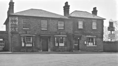 Booth Collection Black and white photo shows pub with windows and chimneys next to a sign for ale and stout