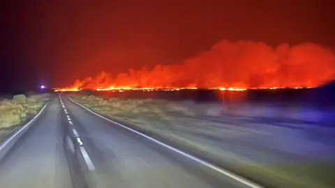 orange flames and billows of smoke in a grassy field with a clear road running alongside it