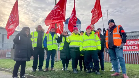 Transport workers on the picket line. A group of men and women are standing on the grass outside their depot in Beeston. Most are wearing high-vis jackets, and some are holding red Unite union flags.