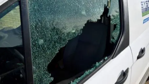 A white van with its window smashed. Another two white vans can be seen parked behind it in a row.