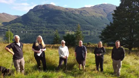 Six people from the Kyle and Lochalsh Community Trust are standing in a field in bright sunshine. The dark waters of Loch Duich are behind them and beyond there are high mountains covering in forestry. 