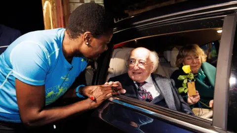PA Media Michael D Higgins and his wife Sabina sit in the back of a car as woman dressed in running clothes talks to them through an open window. Mr Higgins iswearing a suit and a white shirt with a colourful tie. Mrs Higgins is wearing a green dress and holding a yellow rose. The runner talking to them is black and with short dark hair and is wearing a blue running top.