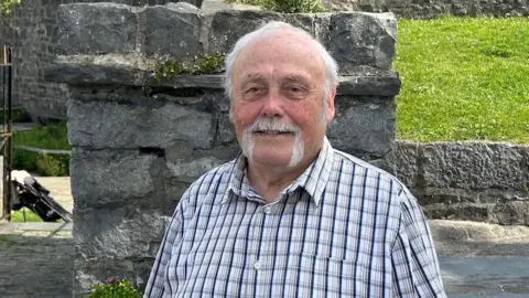 TONY BROWN Tony Brown, who has short white hair, a moustache and a beard, smile. He is wearing a white and purple checked short-sleeve shirt with part of the wall surrounding Castle Rushen behind him next to a grass bank behind him.