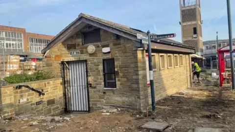 A single story toilet block with a gated doorway and a row of windows along one side