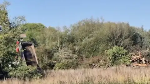 The still image shows blue sky trees in the background and long grass in the foreground. The foreground also contains a dust cart which appears to be tipping material out of the back. 
