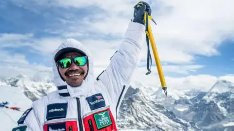 Mr Magar wearing a white coat and holding a yellow pole celebrates reaching the peak of Mount Everest