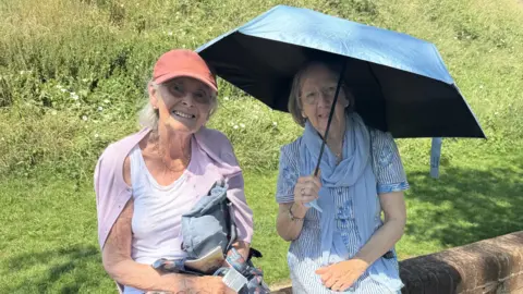 Ronnie Chilcott and Row Hemingway sat on a wall at the bottom of a grass bank. One of the women is holding an umbrella to provide shade.