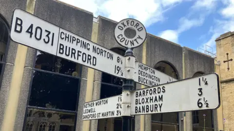 An old-style road sign outside County Hall in Oxford shows the direction and distances to a number of different places in and around Oxfordshire.