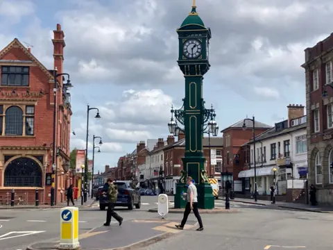 A view of buildings and structures, including a tall green Victorian clock with gilt accents in the centre of a mini roundabout. A red brick three-storey pub with leaded windows is on the left. People cross a road between safety bollards and a car drives down a street.