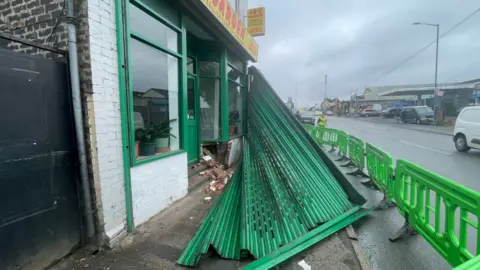 The image is a closer, side-on view of the shop's green metal barrier hanging over the pavement from the impact of the crash. It is still attached to one end of the takeaway. The scene is cut off by green barriers partially in the road.