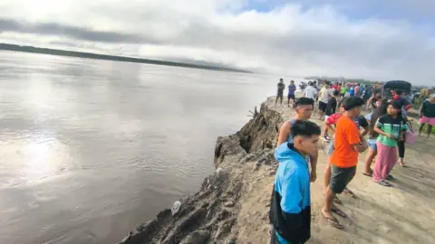 @COENPeru/X A crowd of people stand on the bank of the Ucayali River