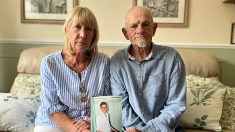 Lesley and Phil Russell hold up a photo of their son. They are sitting on a leather sofa in a living room. Lesley on the left has blonde, shoulder length hair with a fringe. She is wearing a blue striped top and white trousers. Phil is wearing a blue shirt. He has grey hair and a grey goatee beard. The photo they are holding up is of Nik when he was a teenager, he is wearing a white shirt and is smiling at the camera.