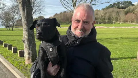 A man wearing a black coat holding his Patterdale Terrier dog, also wearing a black harness, standing by the park with blue skies and trees behind.
