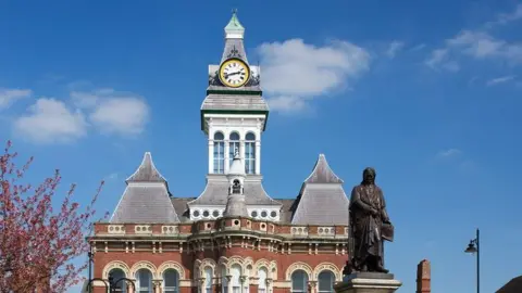 Getty Images The clock tower at Grantham Guildhall, which is mostly white and has a gold and white clock face. It is on top of a bigger building with a grey pointed roof and a statue in front of it.