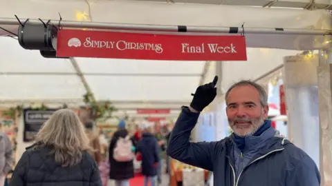 John Garton pointing up to a sign which says "Simply Christmas Final week". He is stood on the right and is wearing a blue coat. Behind him is a gazebo filled with people walking around and looking at stalls. 