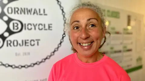 Emma Roberts, with tied-back curly grey hair and wearing a dark pink top smiles at the camera