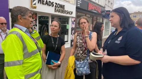 A man in a high-visibility vest speaks to three women in a high street with a barbershop and a barbecue restaurant in the background.