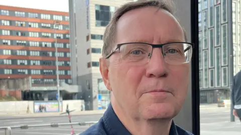 John Myers, who wears glasses and is clean-shaven, is photographed in a dark blue shirt.