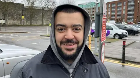 Phil Harrison/BBC A man with a black wearing and short hair wearing a grey hoodie with the hood up under a black coat. They are standing in a car park.