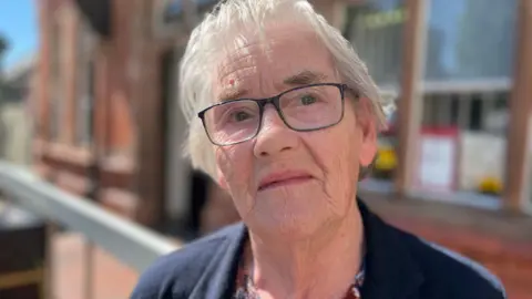 An 86-year-old woman with grey hair wears dark glasses and a dark top is looking straight at the camera.